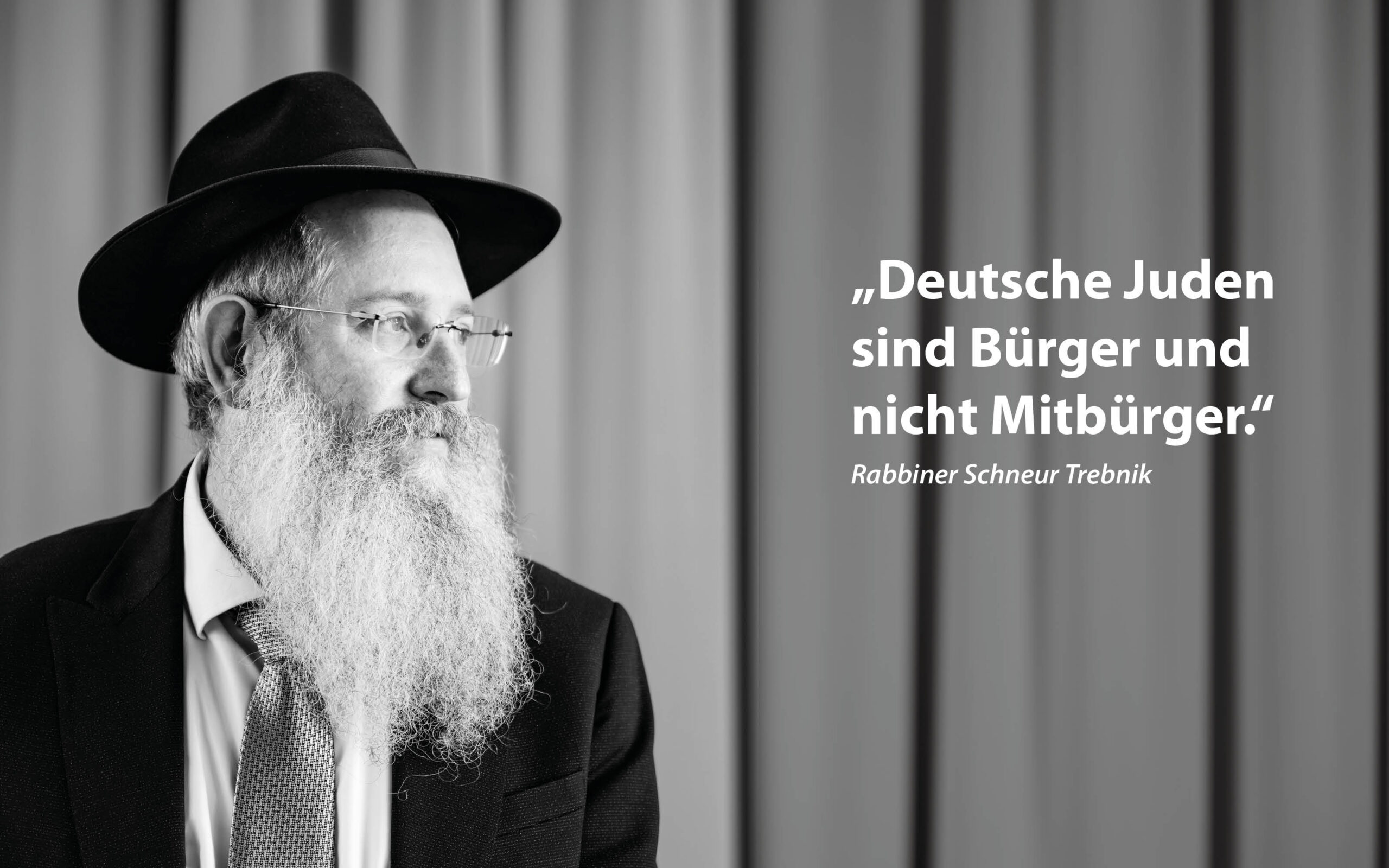 Schwarz-Weiß-Fotografie eines bärtigen Mannes mit Hut, Brille, Anzug und Krawatte. Er blickt nach links. Im Hintergrund sind vertikale Vorhänge zu sehen. Rechts neben ihm steht ein Zitat auf Deutsch: „Deutsche Juden sind Bürger und nicht Mitbürger.“ – Rabbiner Schneur Trebnik.