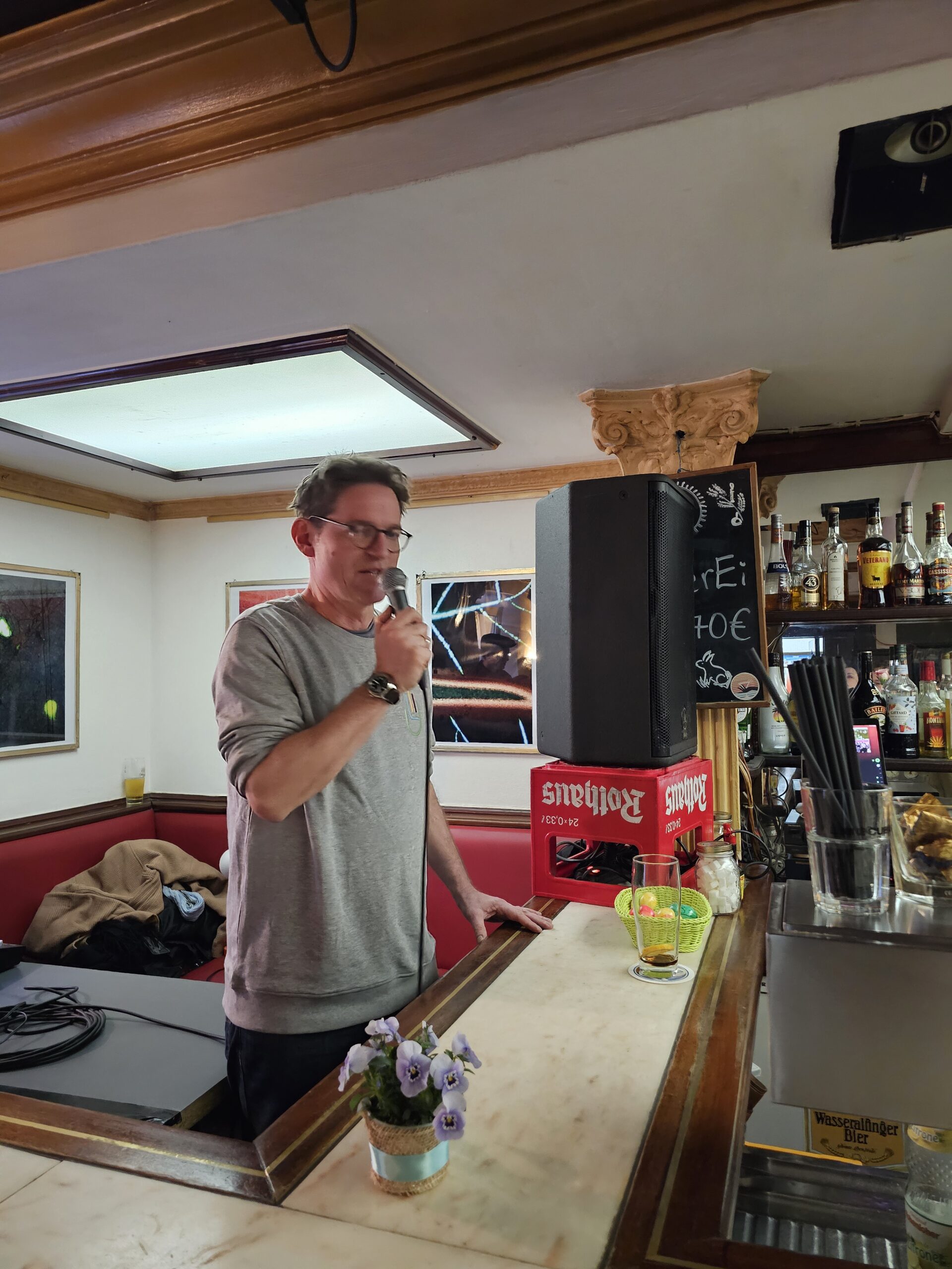 Ein Mann mit Brille und grauem Sweatshirt steht hinter einem Bartresen und hält ein Mikrofon in der Hand. Neben ihm stehen verschiedene Getränke und ein Lautsprecher. Auf dem Tresen steht ein kleiner Blumentopf. Im Hintergrund sind gerahmte Bilder und ein Barregal mit Flaschen zu sehen.