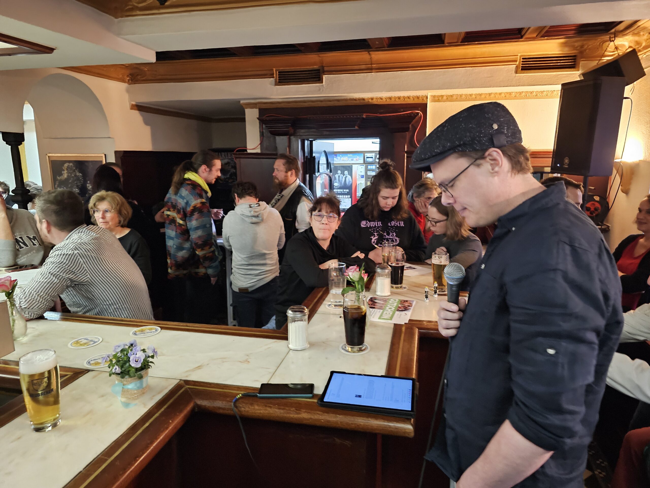 Ein Mann mit einem Mikrofon steht vor einem belebten Pub. Gäste sitzen an der Bar und an den Tischen und unterhalten sich. Getränke stehen auf der Theke und tragen zur lebhaften Atmosphäre bei.