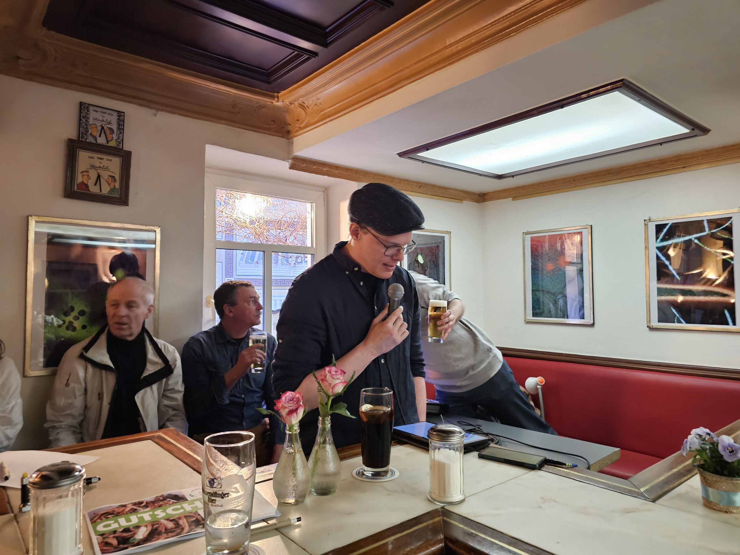 Ein Mann mit schwarzer Mütze und Jacke steht mit einem Drink in der Hand an einer Bar neben einem Mikrofon. Im Hintergrund sitzen drei Männer an einem Tisch. Der Raum ist mit gerahmten Kunstwerken und Blumen auf der Theke dekoriert.