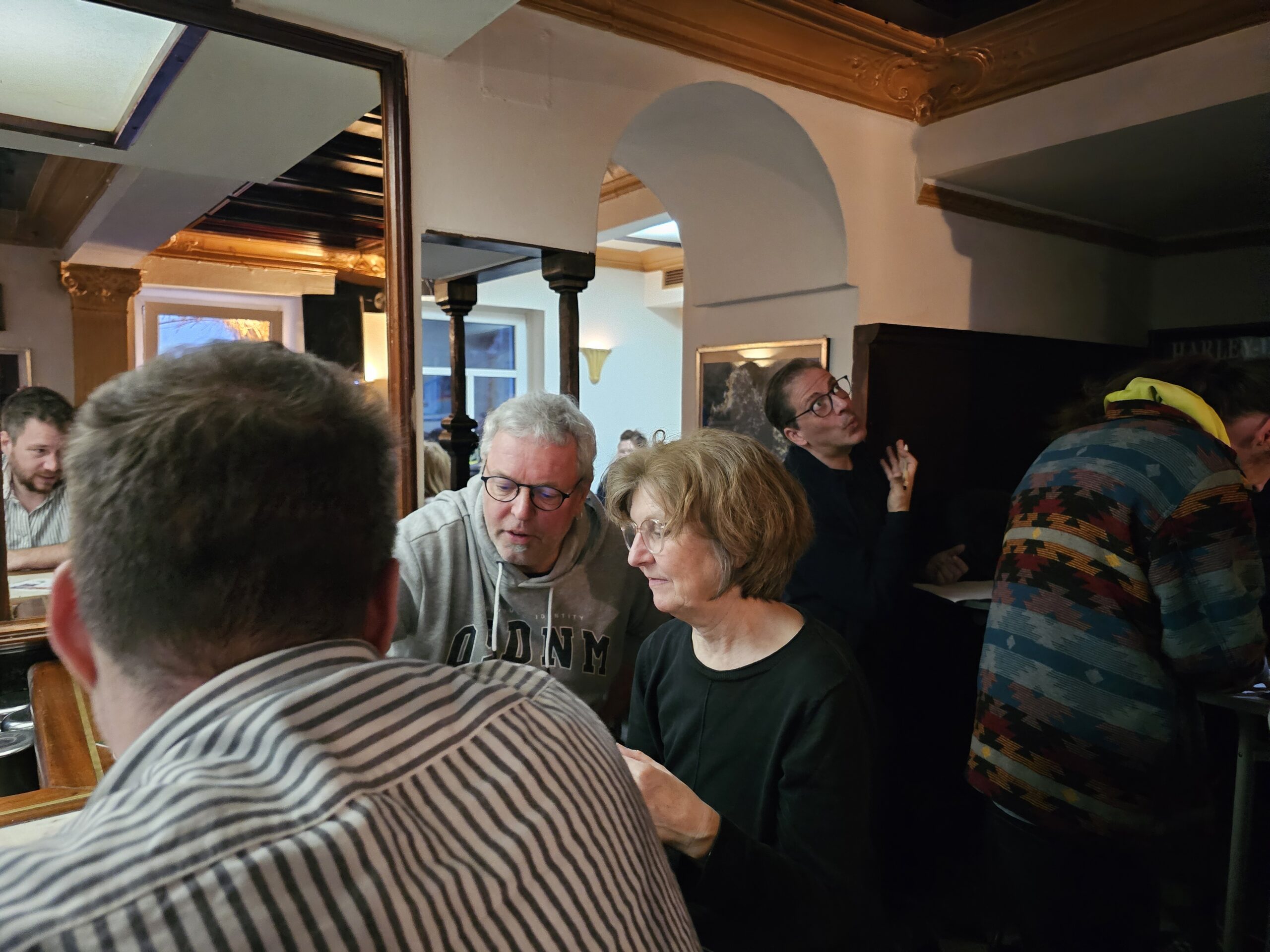 Eine Gruppe von Menschen sitzt in einer gemütlichen Bar. Ein Mann mit grauem Kapuzenpulli und Brille und eine Frau mit kurzen Haaren unterhalten sich an einem Tisch. Andere sitzen und stehen herum und unterhalten sich. Warmes Licht schafft eine einladende Atmosphäre.