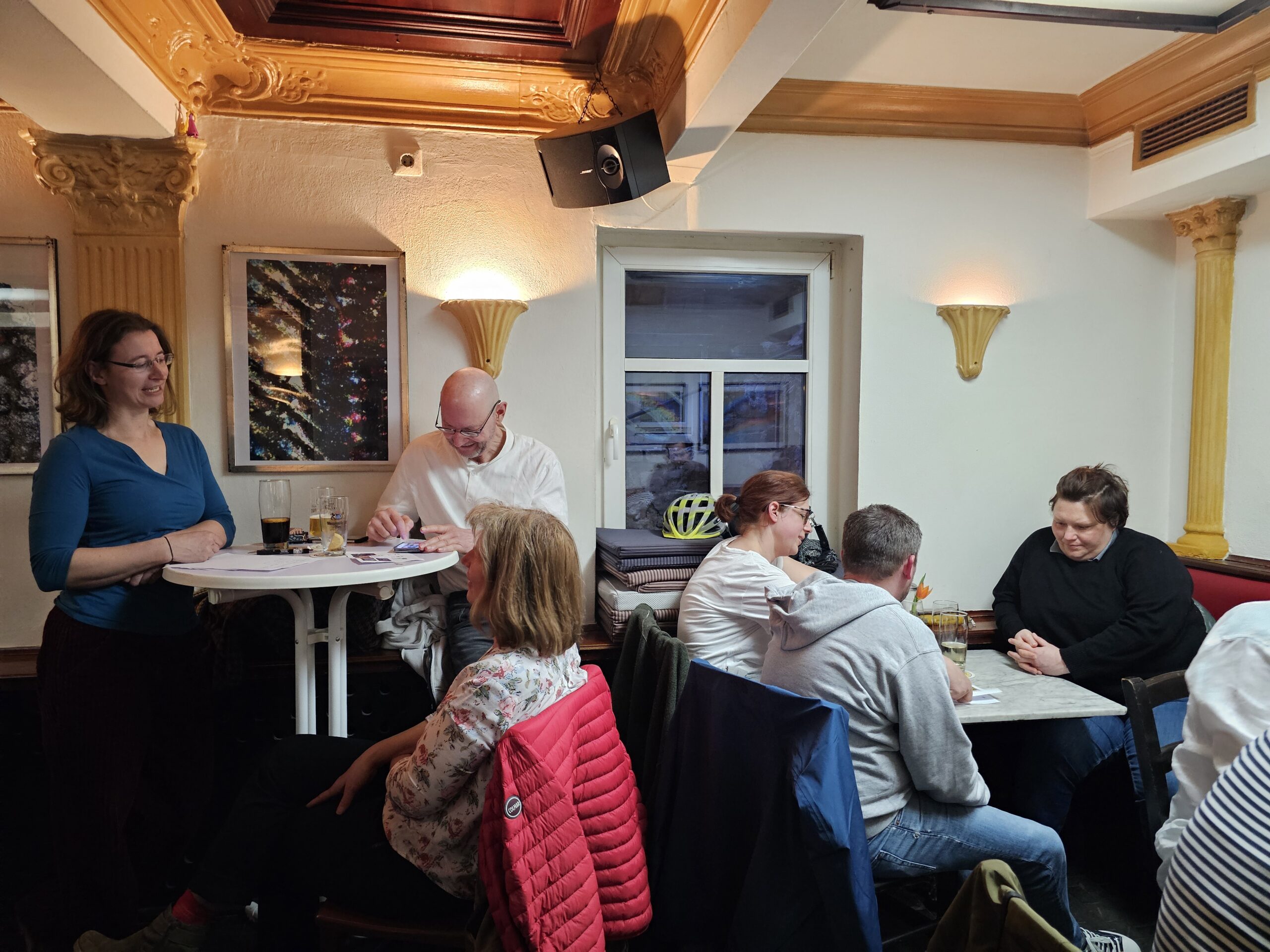 Eine kleine Gruppe von Menschen sitzt an Tischen in einem gemütlichen Café mit warmer Beleuchtung und kunstvollen Deckendetails. Eine Frau steht neben einem hohen Tisch und mehrere Personen unterhalten sich. Auf den Tischen stehen Getränke.