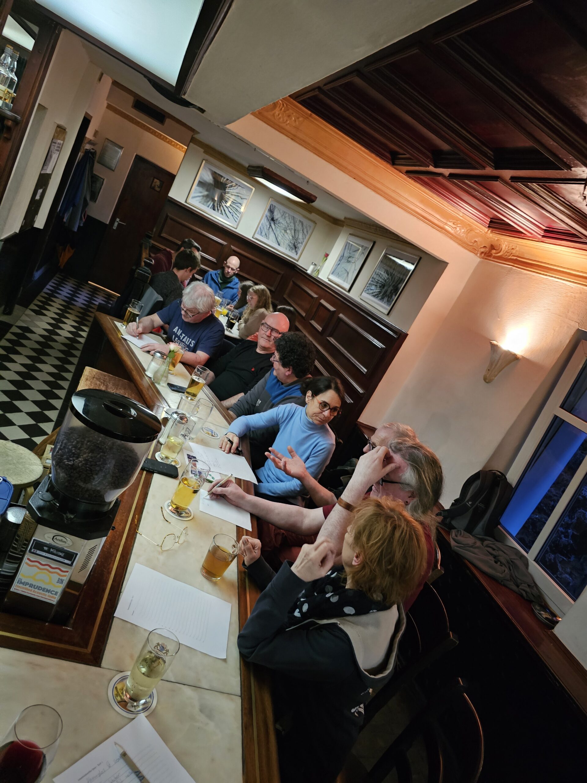 Eine Gruppe von Menschen sitzt in einem Pub um einen rechteckigen Tisch herum, vor ihnen liegen Getränke und Zeitungen. Die Atmosphäre ist warm, mit Holzakzenten und Wandkunst. Der Boden hat ein schwarz-weißes Schachbrettmuster.