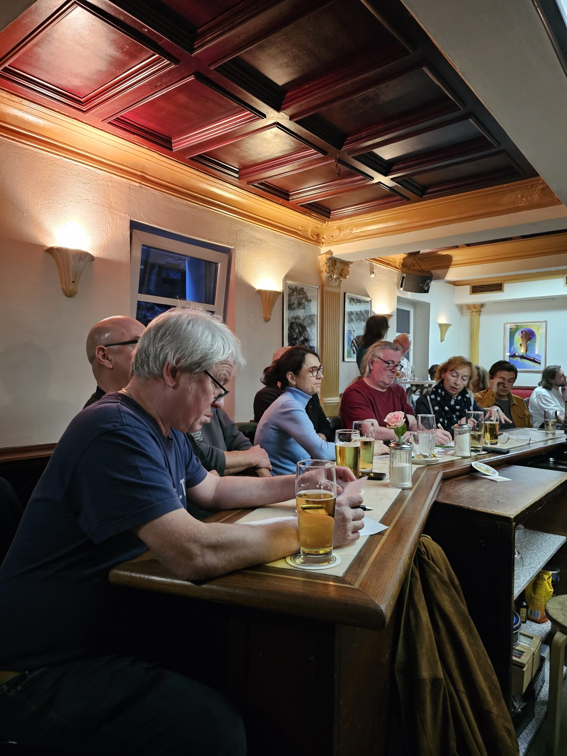 Eine Gruppe von Menschen sitzt an einer Bar, einige haben Biergläser in der Hand und machen sich Notizen auf Papier. Sie sind vertieft, mit einer warm beleuchteten Holzdecke und gerahmten Bildern an cremefarbenen Wänden.