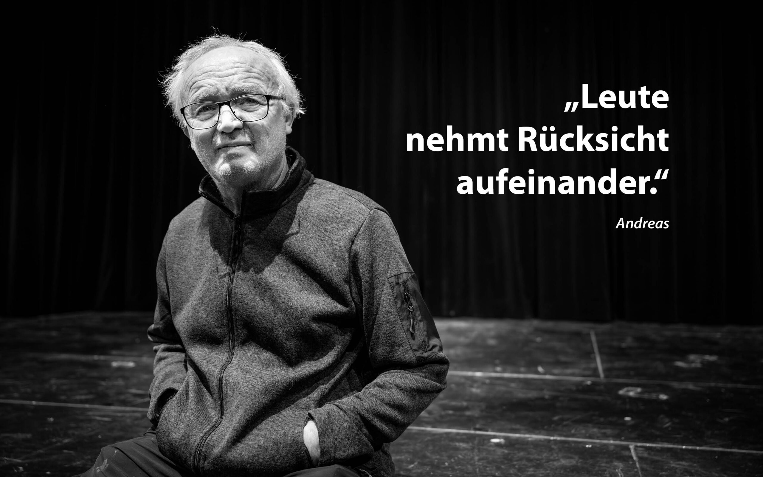 Ein älterer Mann mit Brille und Jacke sitzt auf einer Bühne vor einem dunklen Vorhang. Der deutsche Text neben ihm lautet: „Leute nehmt Rücksicht aufeinander.“ gefolgt von „Andreas