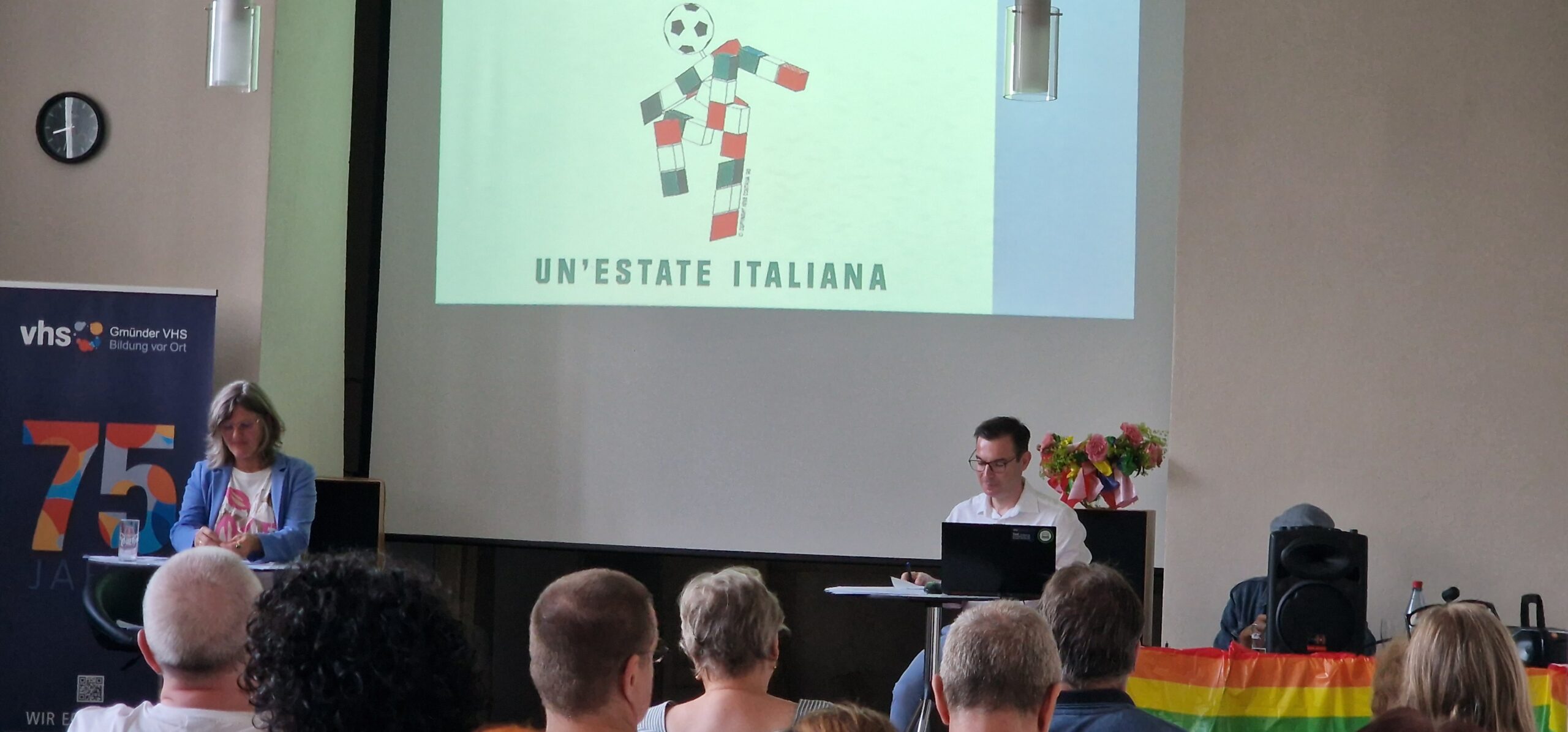 Ein Redner steht an einem Podium, während ein Publikum zuhört. Auf einem Bildschirm erscheint der Text „Un'estate Italiana“ mit einem stilisierten Fußballspieler aus bunten Blöcken. Über einem Tisch hängt eine Regenbogenfahne. Eine andere Person sitzt an einem Laptop.