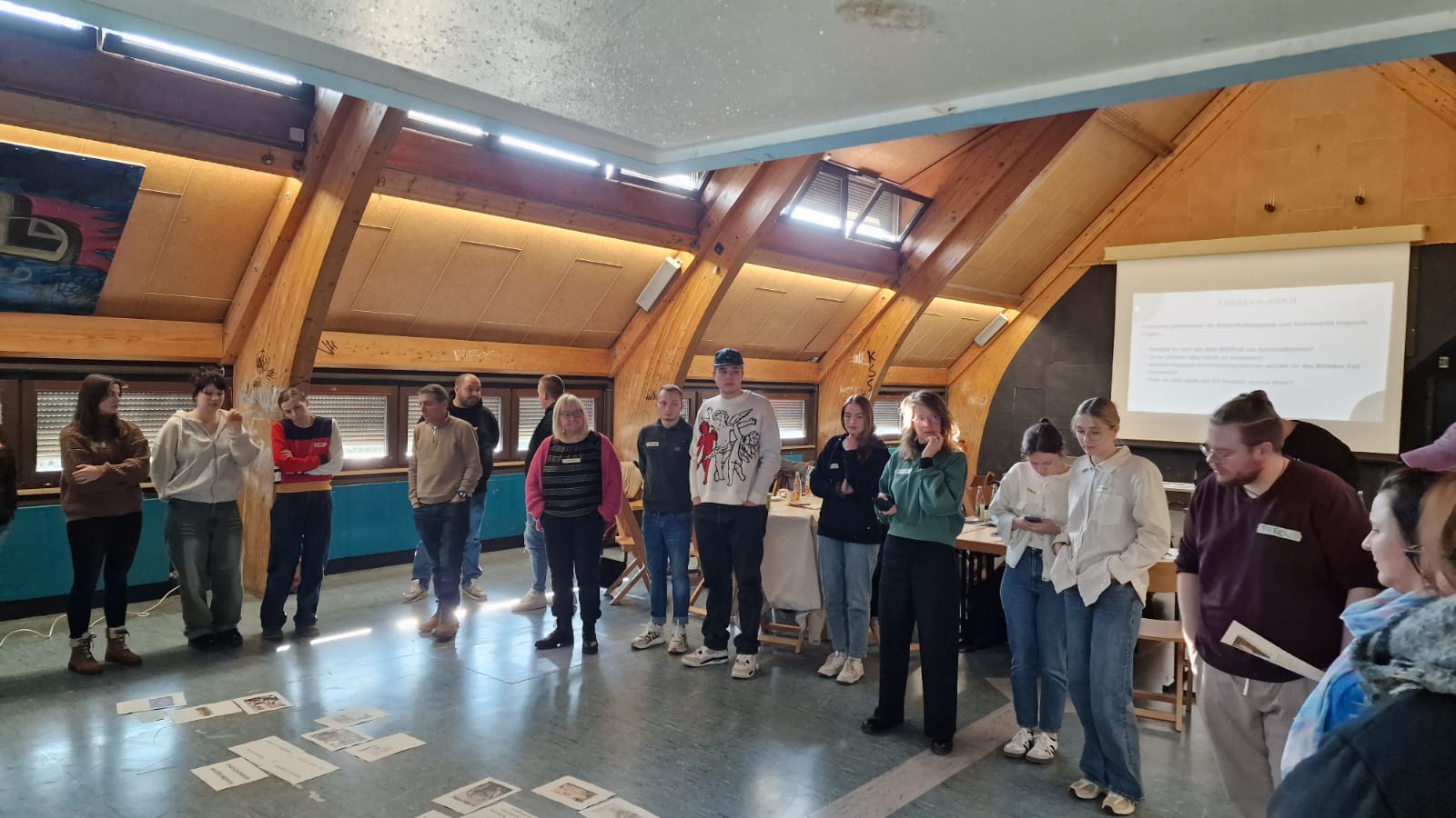 Eine Gruppe von etwa 17 Personen steht im Halbkreis in einem Raum mit Holzdecke und großen Fenstern. Der Raum wirkt wie ein Seminar- oder Gruppenraum. Auf dem Boden liegen verschiedene Zettel mit Texten und Bildern. Im Hintergrund ist ein Beamerbild zu sehen mit einer Präsentationsfolie. Die Teilnehmenden schauen nachdenklich auf das, was am Boden ausgelegt ist. Die Atmosphäre wirkt konzentriert und ruhig.