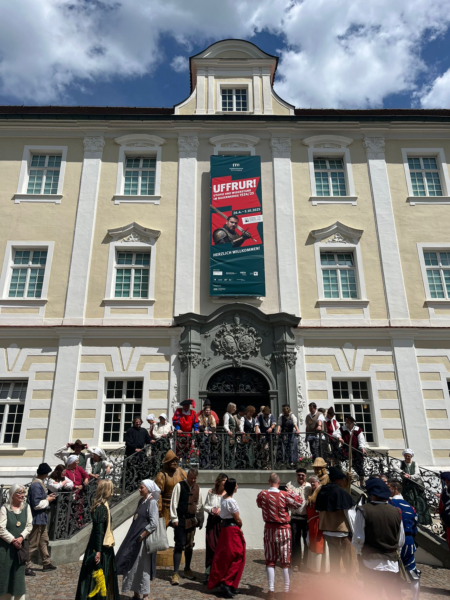 Vor einem großen, hellgelben Barockgebäude mit weißen Fensterrahmen und einem prunkvoll verzierten Portal haben sich zahlreiche Menschen in historischen Kostümen versammelt. Einige stehen auf der Treppe und dem Podest vor dem Eingang, andere unten auf dem gepflasterten Platz. Die Kleidung erinnert an die Zeit des 16. Jahrhunderts: Männer tragen Wämser, Hüte und weite Hosen, Frauen tragen lange Kleider, Schürzen und Kopftücher. Über dem Portal hängt ein großes grünes Banner mit rotem Motiv und dem Titel: „UFFRUR! Utopie und Widerstand im Bauernkrieg 1524/25“. Darunter steht: „Herzlich willkommen!“ Der Himmel ist teils bewölkt, und Sonnenlicht fällt auf die Szene. Die Stimmung wirkt lebendig und festlich, als bereite sich die Gruppe auf eine Führung oder ein gemeinsames Programm vor.