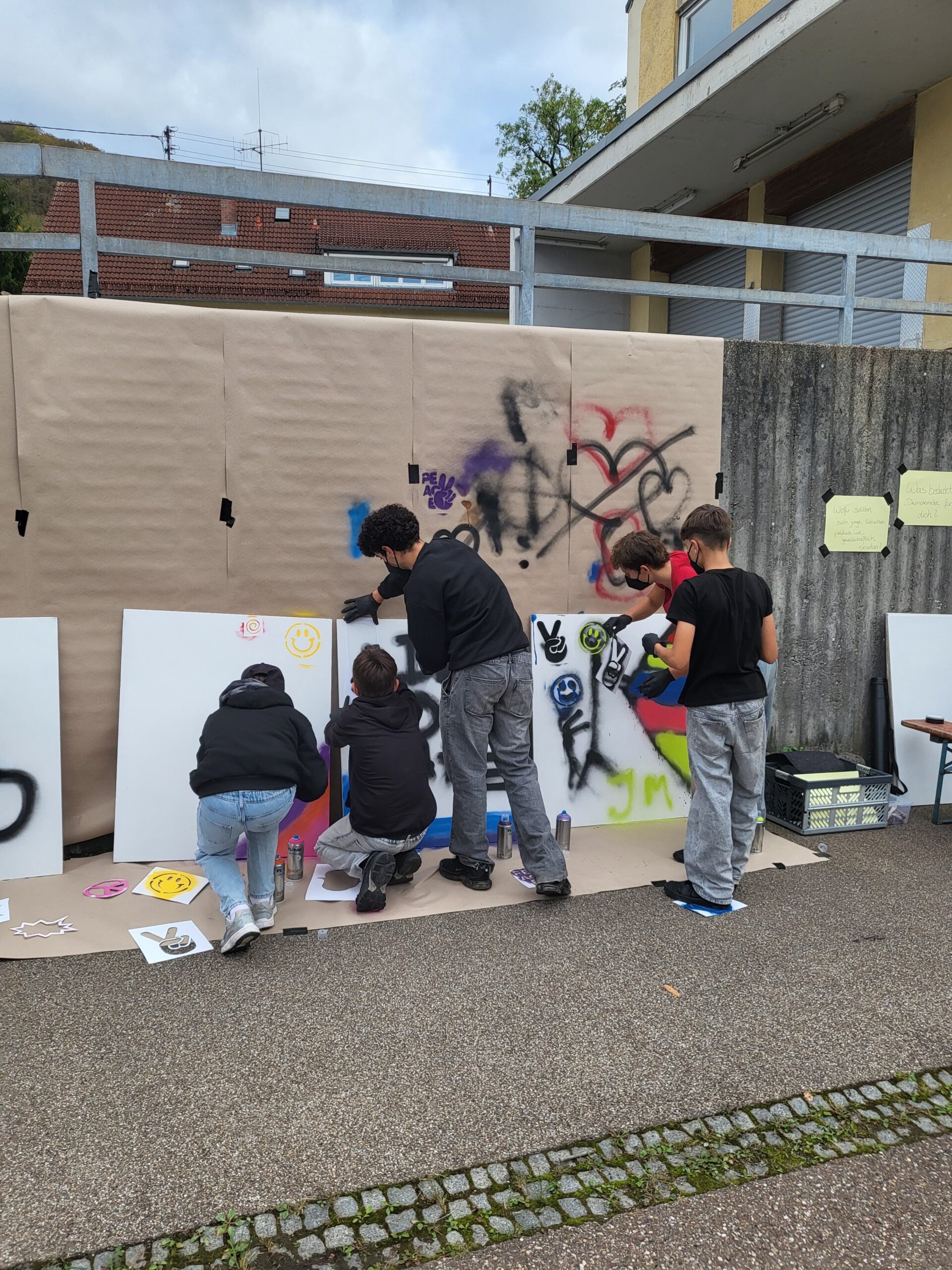 Fünf Jugendliche gestalten im Freien große weiße Bildtafeln mit Sprühfarben und Schablonen. Dabei entstehen bunte Motive, darunter lächelnde Gesichter und Friedenssymbole.