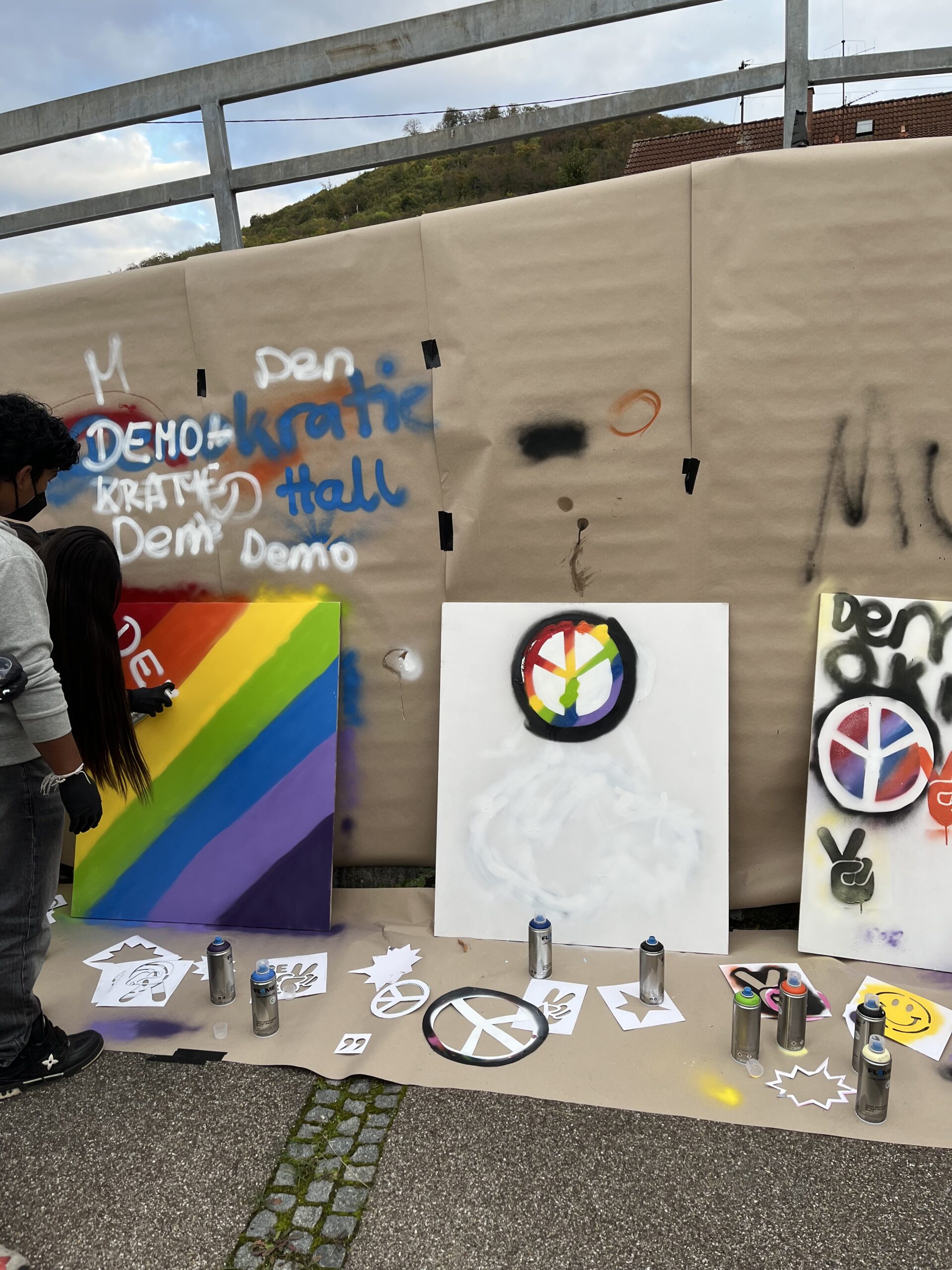Mehrere Graffiti-Bilder zeigen Regenbogenstreifen und bunte Friedenszeichen. Vor den Bildern befinden sich Sprühdosen und Schablonen.