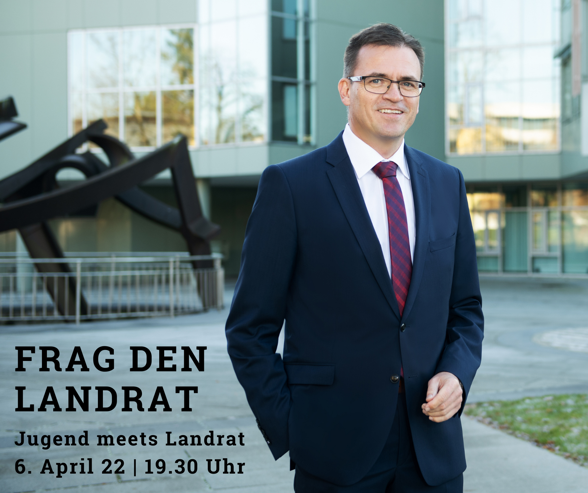 Ein Mann in Anzug und Krawatte steht im Freien vor einem modernen Gebäude. Text auf dem Bild: „FRAG DEN LANDRAT – Jugend trifft Landrat – 6. April 22 | 19.30 Uhr.“.