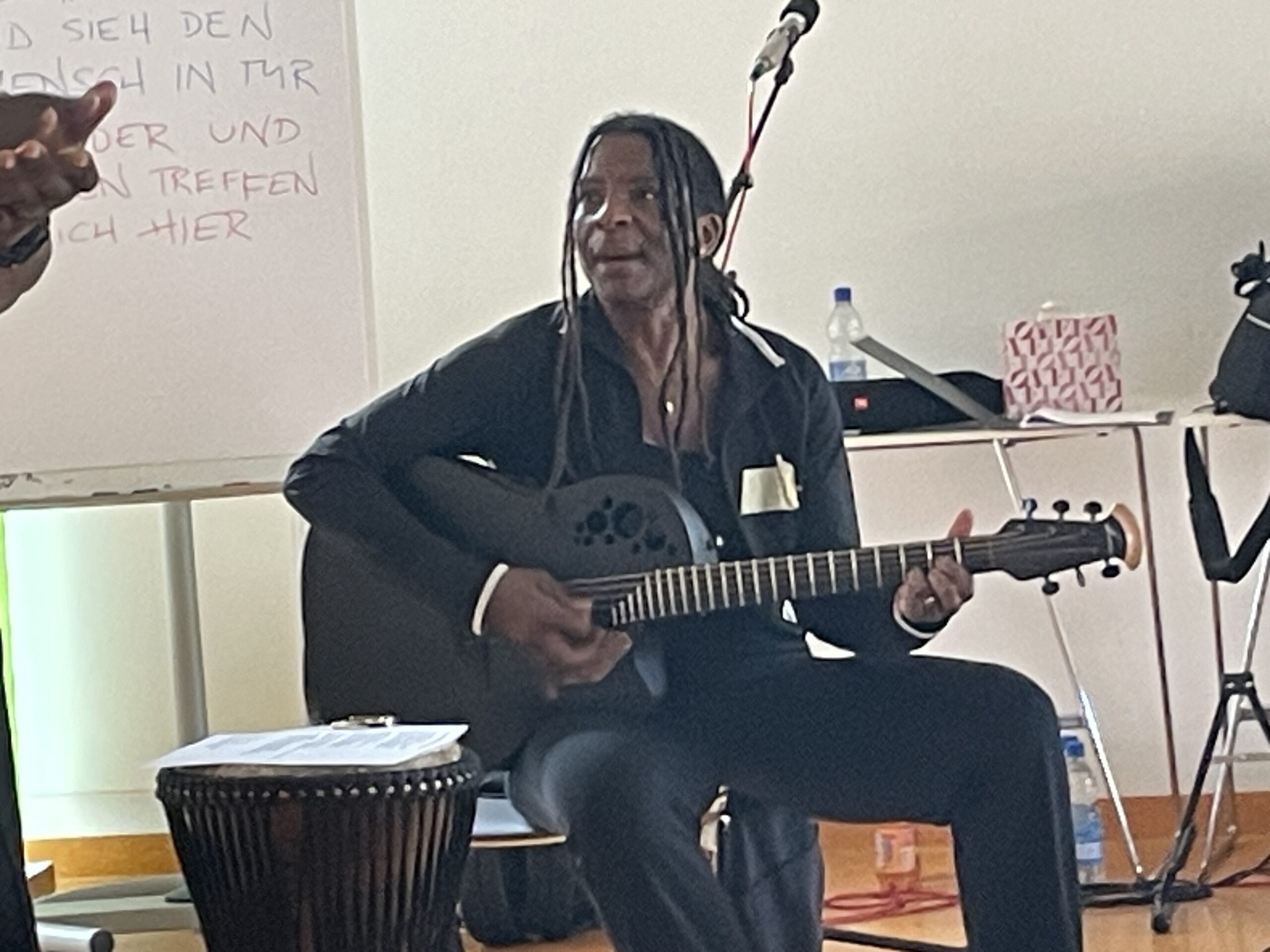 Eine Musikerin sitzt mit einer schwarzen Akustikgitarre vor einem Mikrofon und singt. Vor ihr steht eine Djembe-Trommel. Im Hintergrund sind ein Whiteboard und Veranstaltungsmaterial zu sehen.