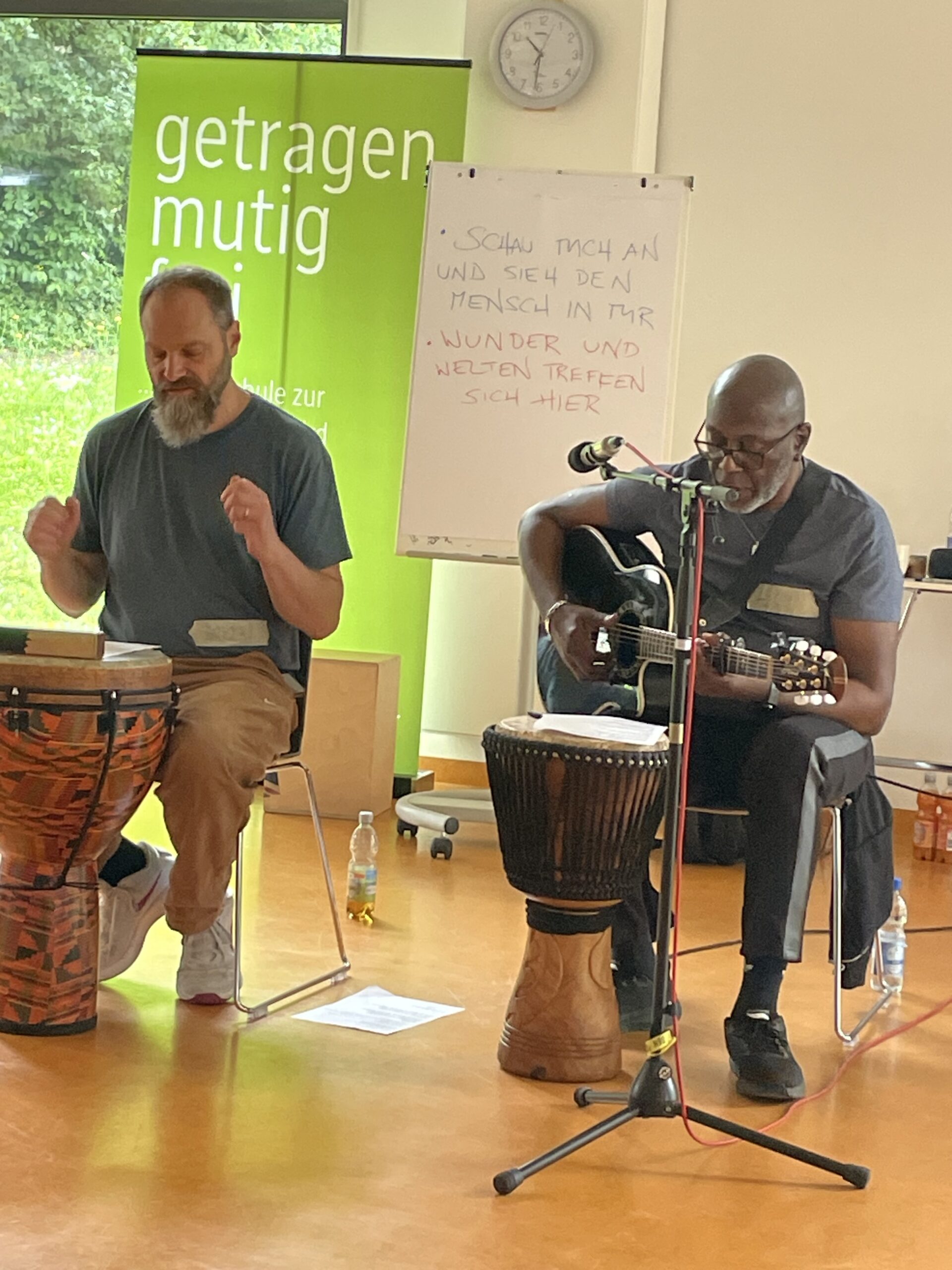 Zwei Musiker gestalten gemeinsam einen Workshop. Eine Person spielt Gitarre und singt, die andere begleitet mit einer Djembe-Trommel. Im Hintergrund stehen ein Whiteboard und ein grünes Banner.