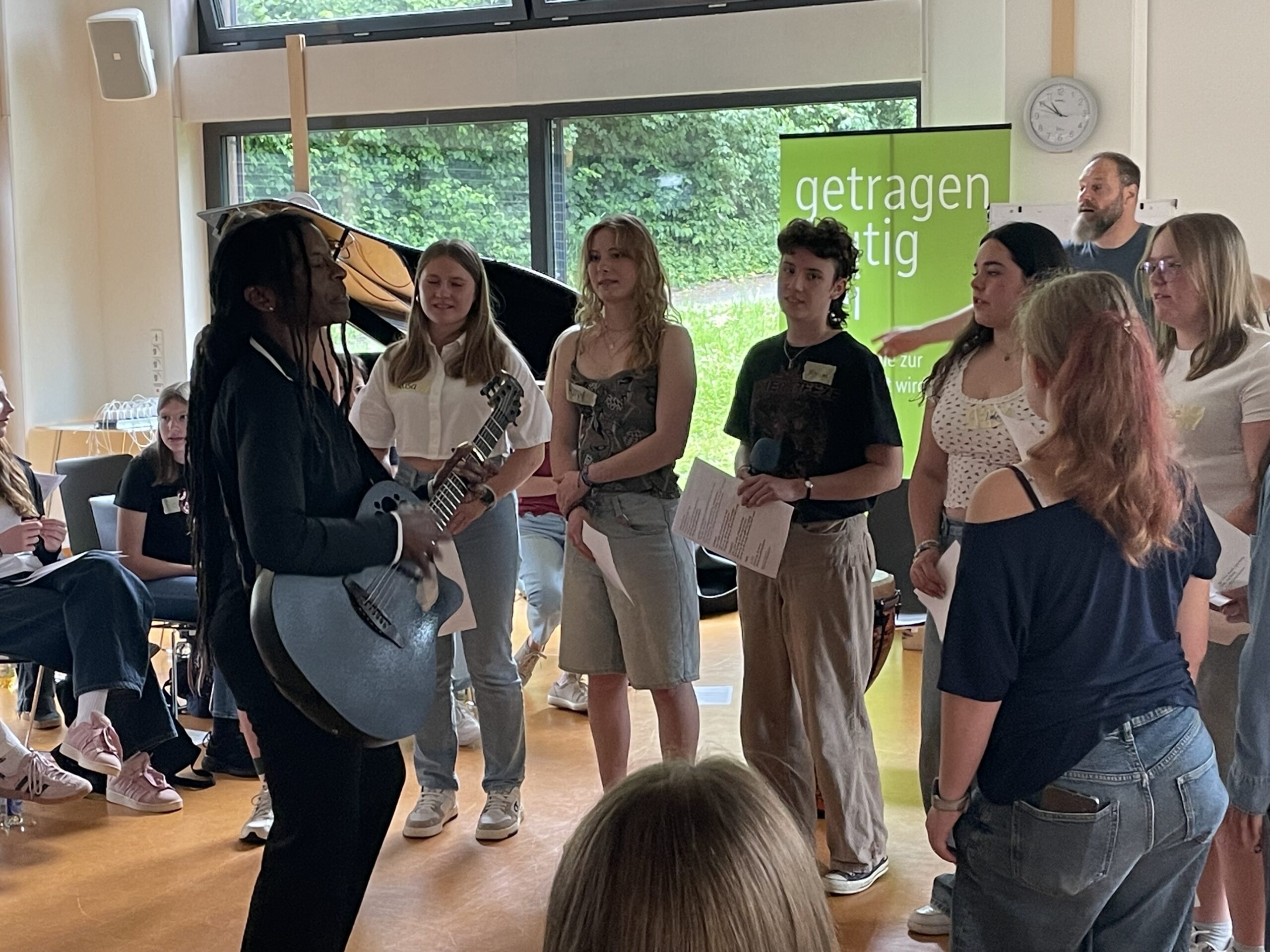 Eine Musikerin steht mit einer Gitarre im Mittelpunkt eines Kreises aus Jugendlichen. Die Teilnehmenden halten Liedblätter in den Händen und singen gemeinsam. Die Szene zeigt einen interaktiven Musikworkshop, bei dem gemeinsames Musizieren und aktives Mitmachen im Vordergrund stehen.