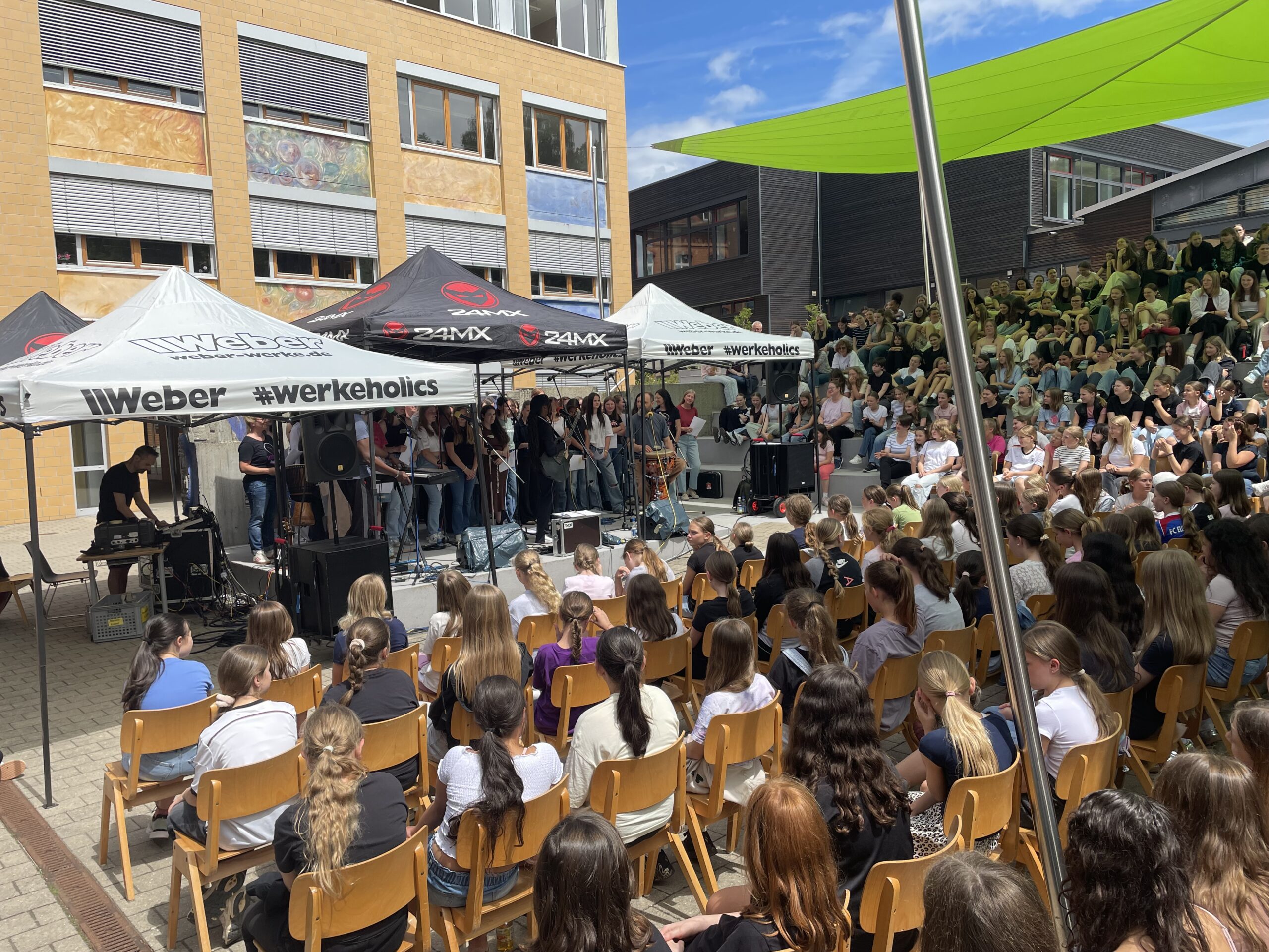 Auf dem Schulhof einer Schule findet eine musikalische Veranstaltung statt. Auf einer Bühne stehen eine Musikerin, ein Trommler sowie ein großer Schülerchor. Vor der Bühne sitzen zahlreiche Schülerinnen und Schüler auf Stühlen, weitere verfolgen die Aufführung von den Treppen des Schulhofs. Die Veranstaltung vermittelt eine gemeinschaftliche Atmosphäre.