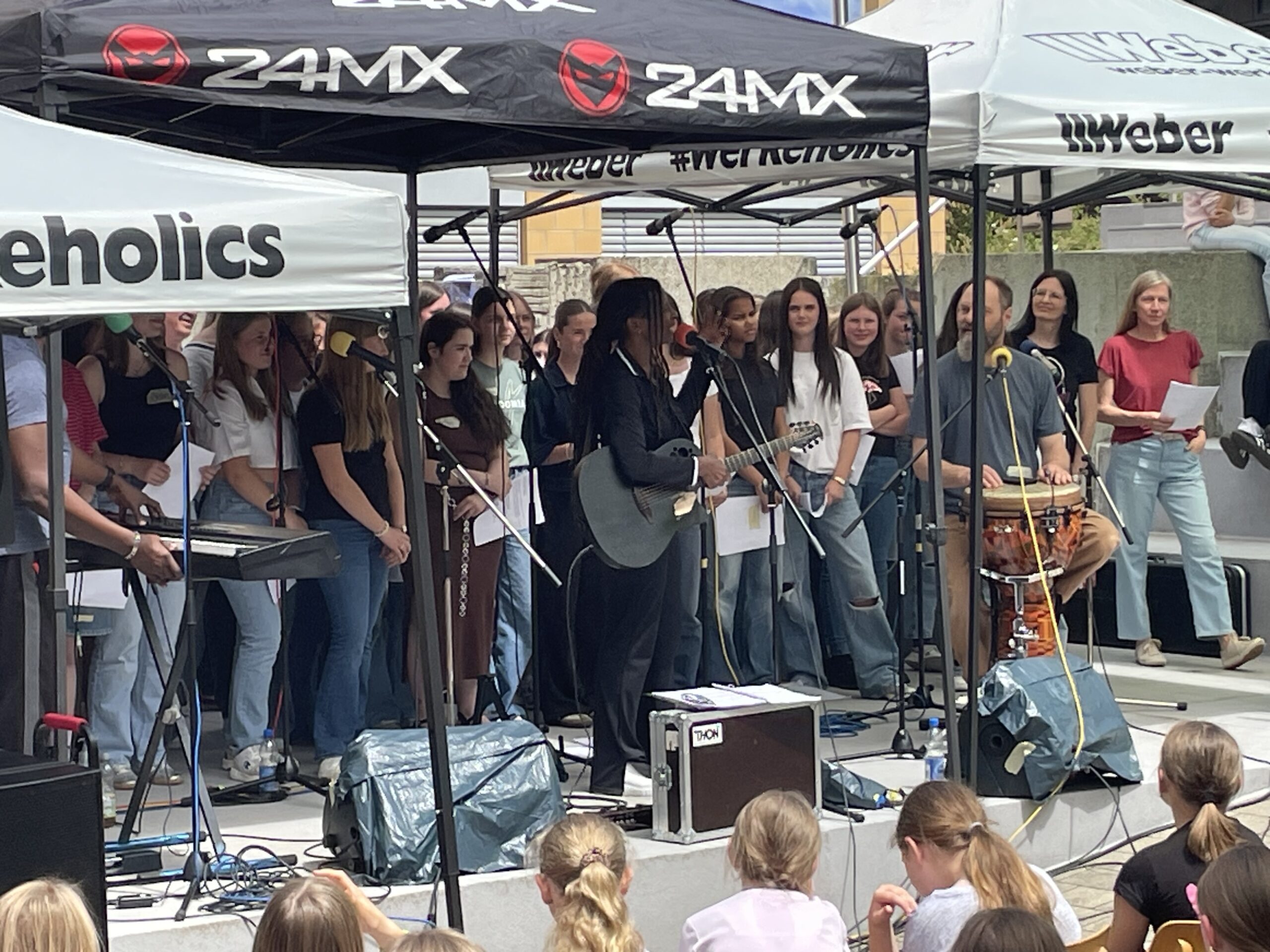 Musikerin mit Gitarre und ein Trommler stehen gemeinsam mit einem Schülerchor auf einer Bühne im Schulhof. Zahlreiche Schülerinnen und Schüler verfolgen den Auftritt.
