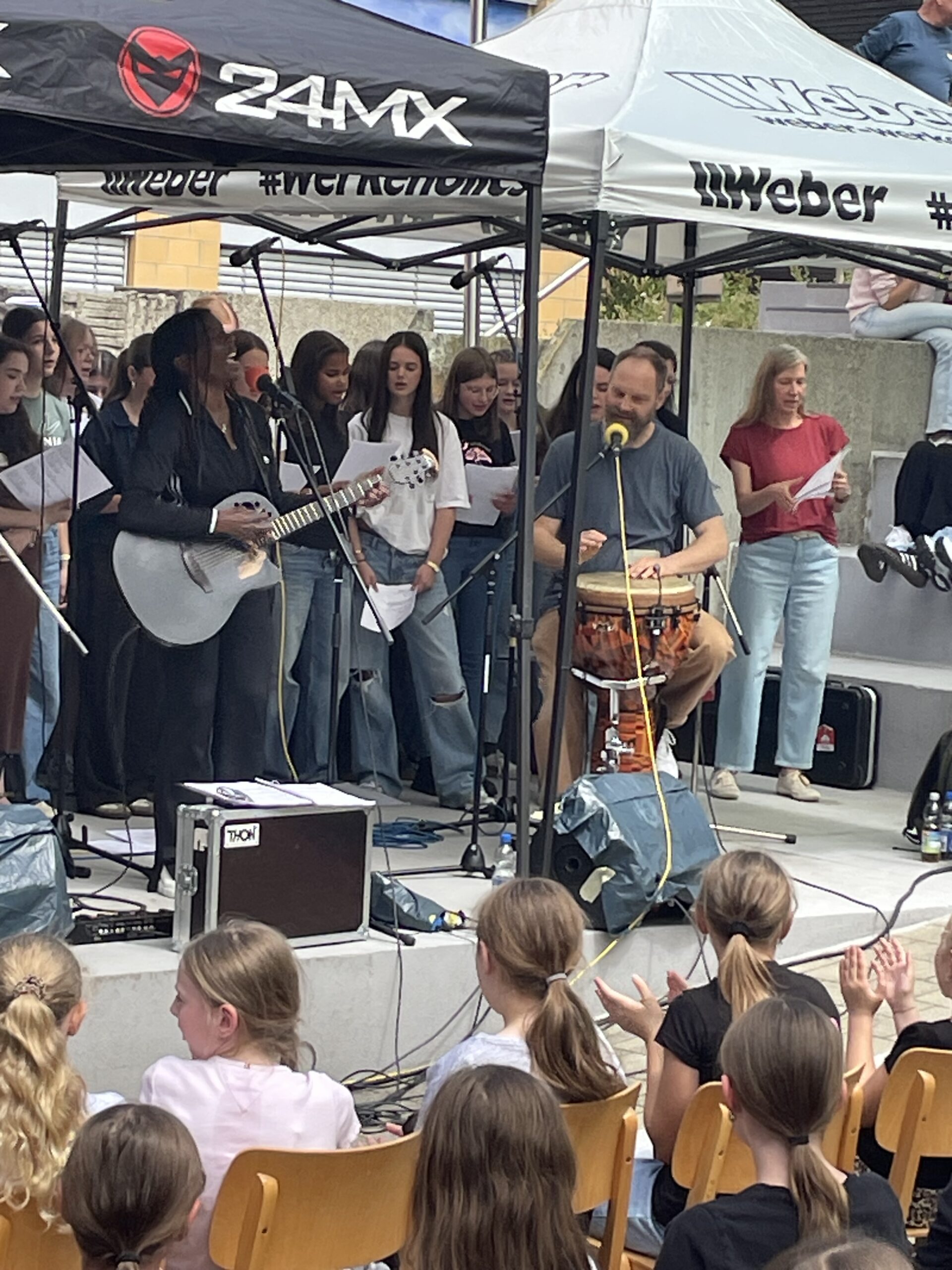 Musikerin mit Gitarre und ein Trommler stehen gemeinsam mit einem Schülerchor auf einer Bühne im Schulhof. Zahlreiche Schülerinnen und Schüler verfolgen den Auftritt.