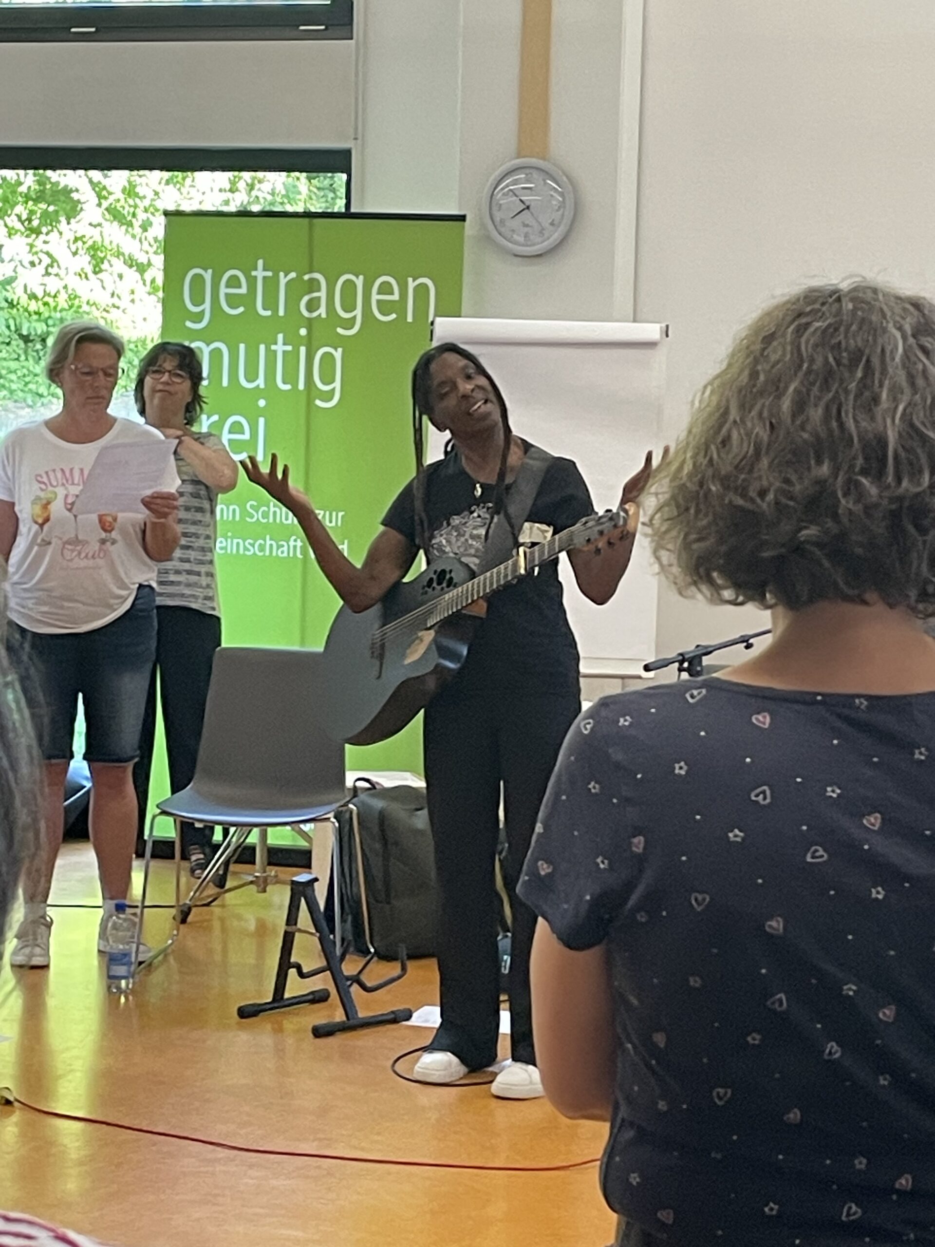 Eine Musikerin singt mit Gitarre vor einer Gruppe von Teilnehmenden in einem Veranstaltungsraum. Zwei weitere Personen begleiten den Gesang mit Liedblättern im Hintergrund.