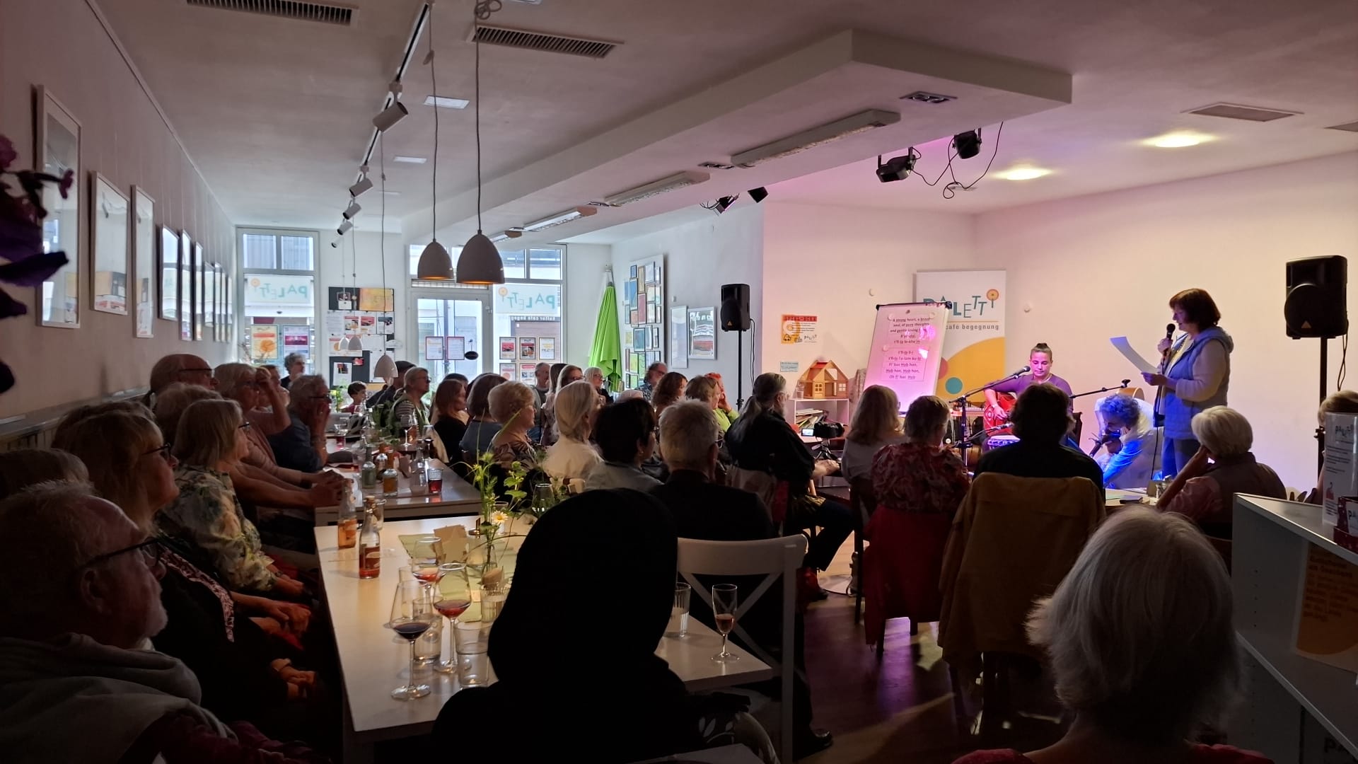 Das Foto zeigt eine Veranstaltung in einem hellen Caféraum, der gut gefüllt ist. Viele Menschen sitzen an Tischen, die mit Gläsern, Flaschen und kleinen Blumen dekoriert sind, und hören aufmerksam zu. Im Hintergrund stehen zwei Frauen auf einer kleinen Bühne: Eine Frau rechts liest aus einem Zettel vor. Die andere Frau (links hinter Mikrofonen) sitzt und spielt ein Instrument, vermutlich eine Trommel. Hinter ihnen ist ein großes, farbiges Banner mit der Aufschrift „PALETTI – Café Begegnung“ zu sehen. Neben der Bühne steht ein Whiteboard oder Flipchart mit Texten darauf. Im Raum herrscht gedämpftes Licht, über der Bühne leuchten Spots in Lila und Rosa. Das Publikum wirkt konzentriert und ruhig. Viele Zuhörerinnen und Zuhörer schauen zur Bühne. Durch die großen Fenster im Hintergrund fällt etwas Tageslicht in den Raum.