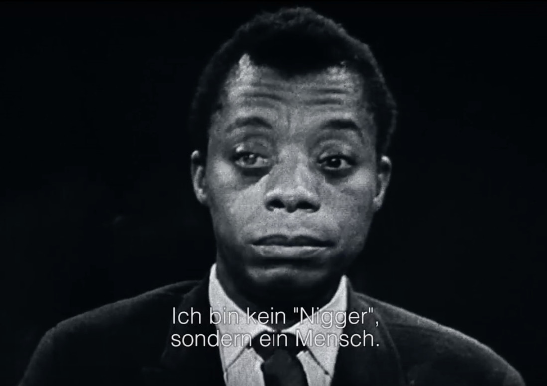 Schwarzweißbild von James Baldwin in Anzug und Krawatte, der mit ernstem Gesichtsausdruck nach vorne blickt. Der deutsche Text unten lautet: „Ich bin kein ‚Nigger‘, sondern ein Mensch.“