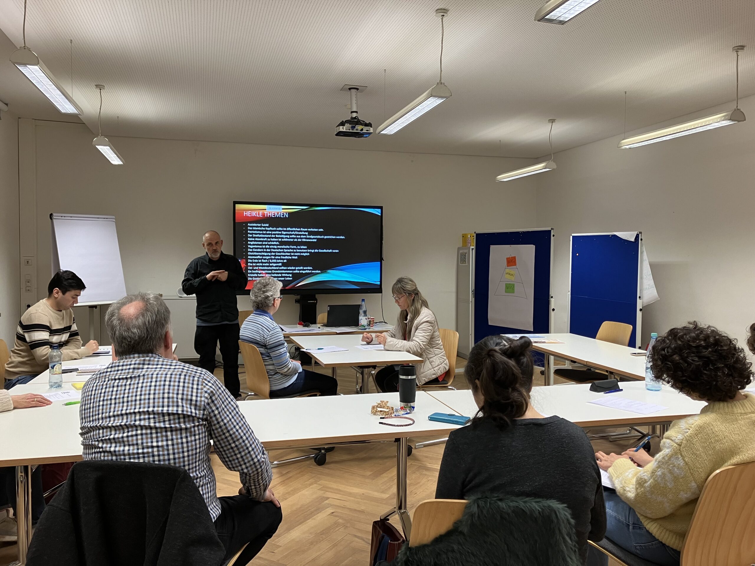 Eine Gruppe von Menschen sitzt in einem Konferenzraum um einen U-förmigen Tisch. Eine Person steht vor einem großen Bildschirm, auf dem eine Präsentation läuft. Auf den Tischen liegen Papiere und Notizbücher. Der Raum ist durch Deckenleuchten gut beleuchtet.