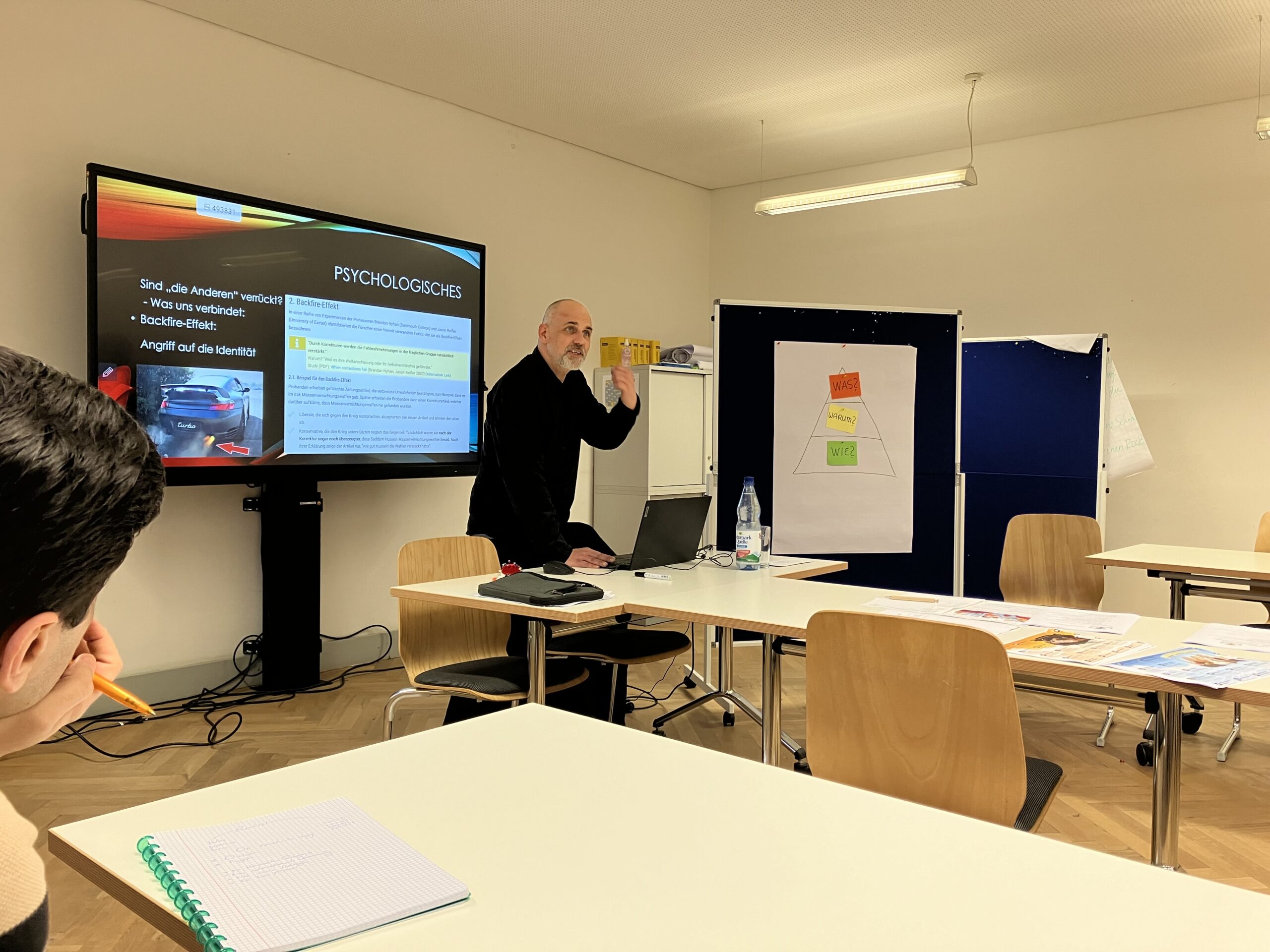 Eine Person steht vor einem Klassenraum und zeigt auf einen großen Bildschirm, auf dem eine Präsentation zu psychologischen Themen läuft. Daneben hängt ein Diagramm auf einem Flipchart. Das Publikum sitzt an den Tischen und macht sich Notizen.