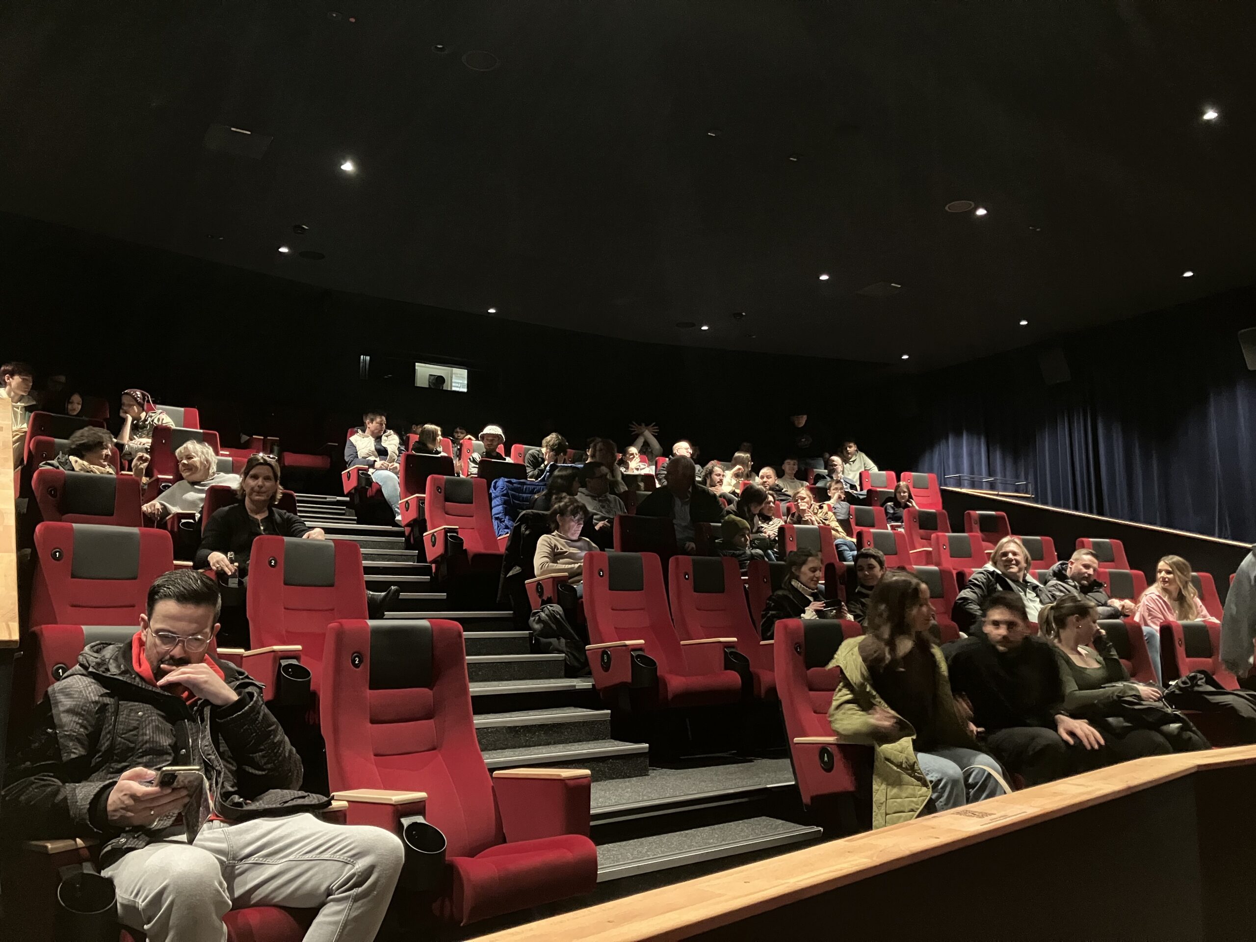 Ein schwach beleuchteter Theatersaal mit mehreren Reihen besetzter roter Sitze. Die Leute scheinen darauf zu warten, dass die Vorstellung beginnt, einige sind mit ihren Handys beschäftigt oder unterhalten sich. Die Stimmung ist entspannt mit sanftem Licht.